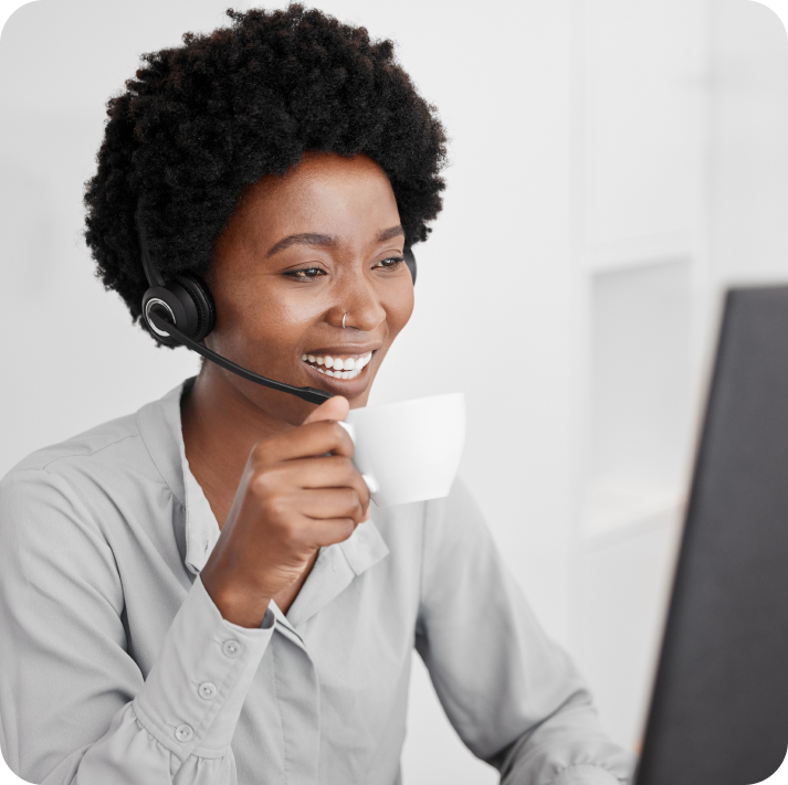 Modisoft female customer support agent sitting in front of a laptop is holding a cup of coffee while talking to a customer via headset.