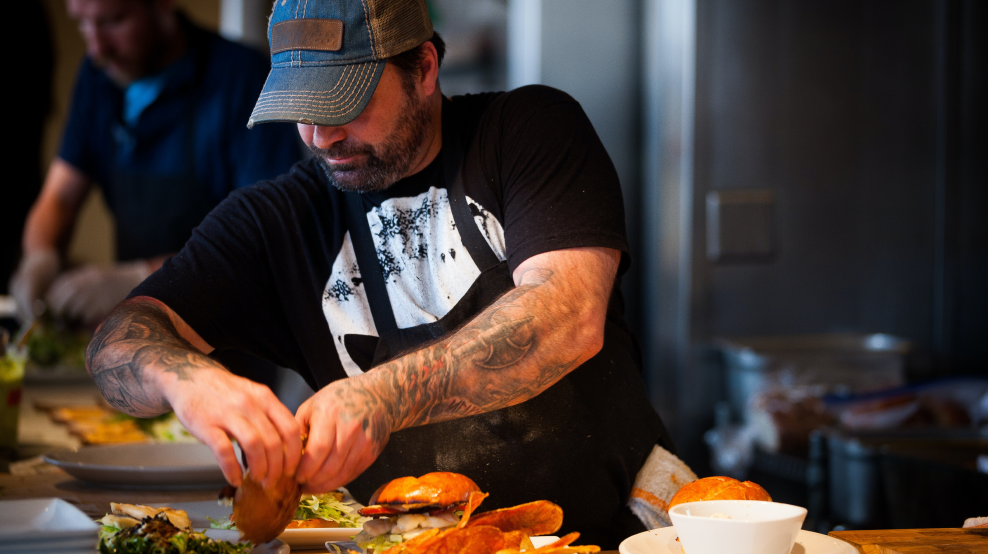 A chef is dressing a burger in the kitchen.