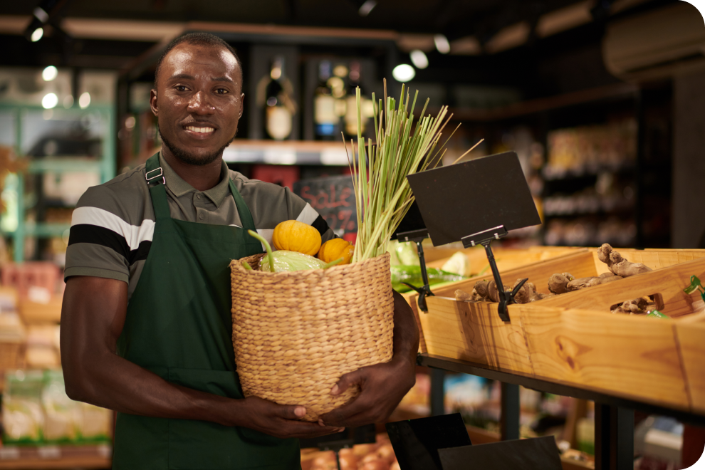 A grocery store employee standing in the grocery section is holding a basket full of groceries.